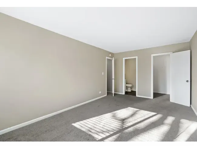Empty room with gray carpet, beige walls, and white doors. Sunlight streams across the floor at Townhomes at Highcrest in Woodridge, IL.