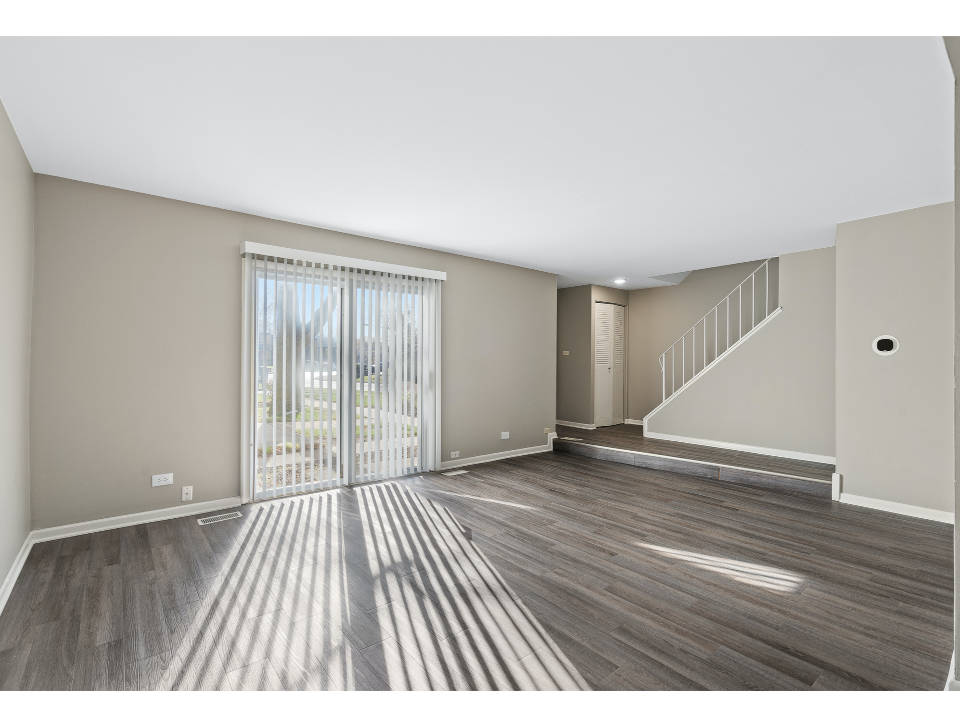 Living room with beige walls, wood-look flooring, sliding glass door with vertical blinds, and a staircase.