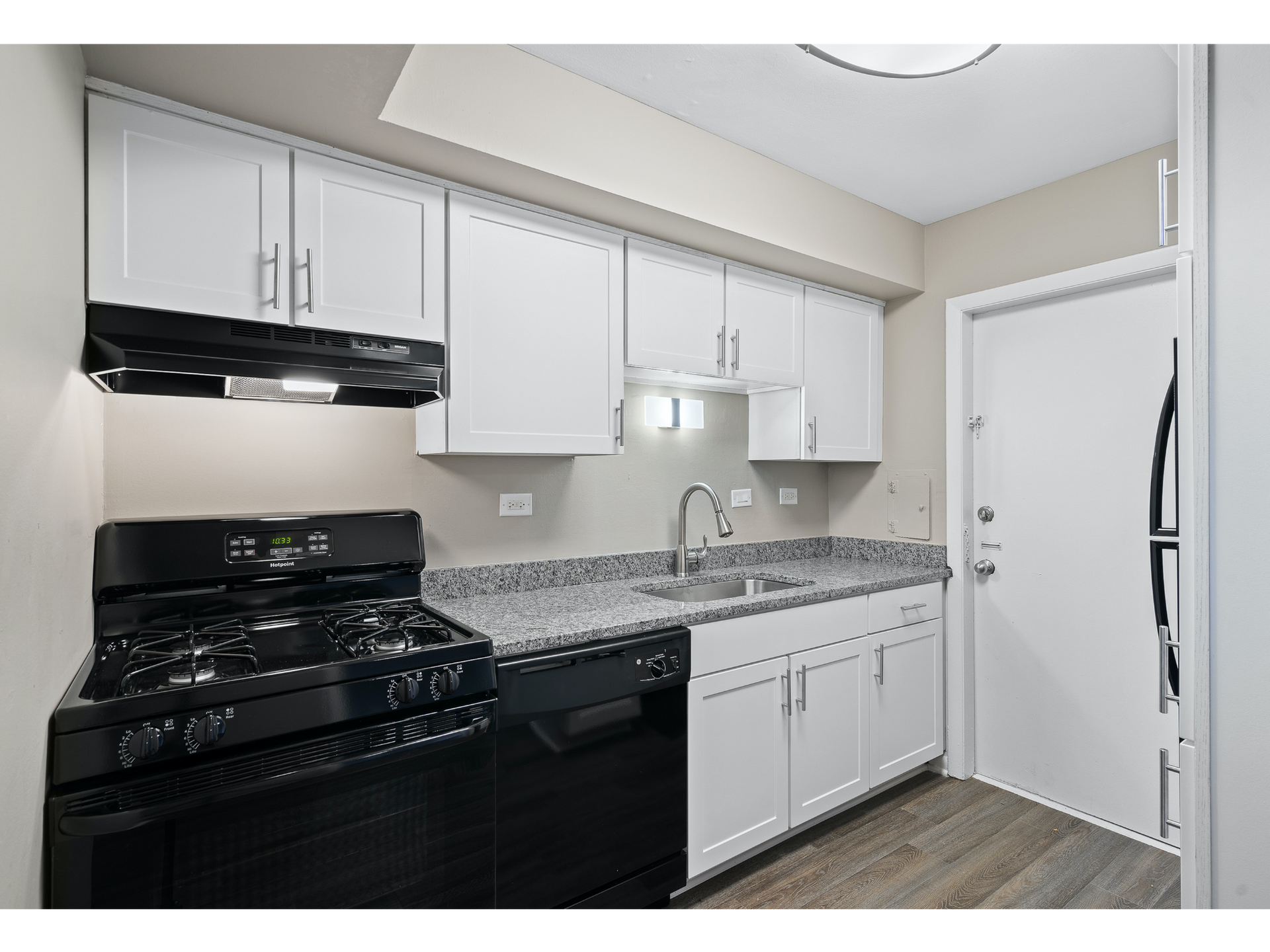 White kitchen with upper and lower cabinets, black stove, stainless sink, and granite counters.