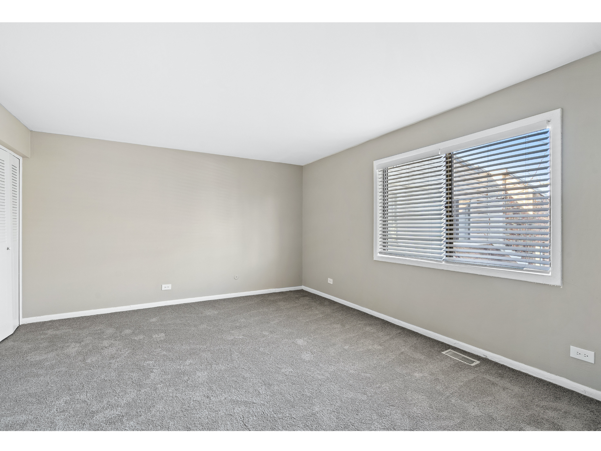 Empty beige bedroom with carpet, a window with white blinds, and a white closet door.