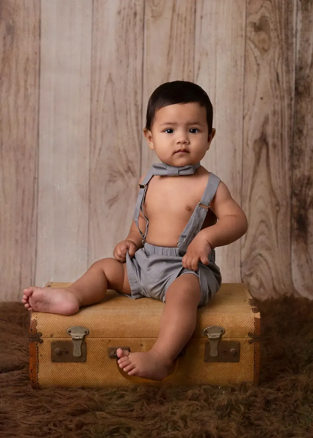 First birthday portrait. Boy seated on old suitcase in shorts and suspenders.