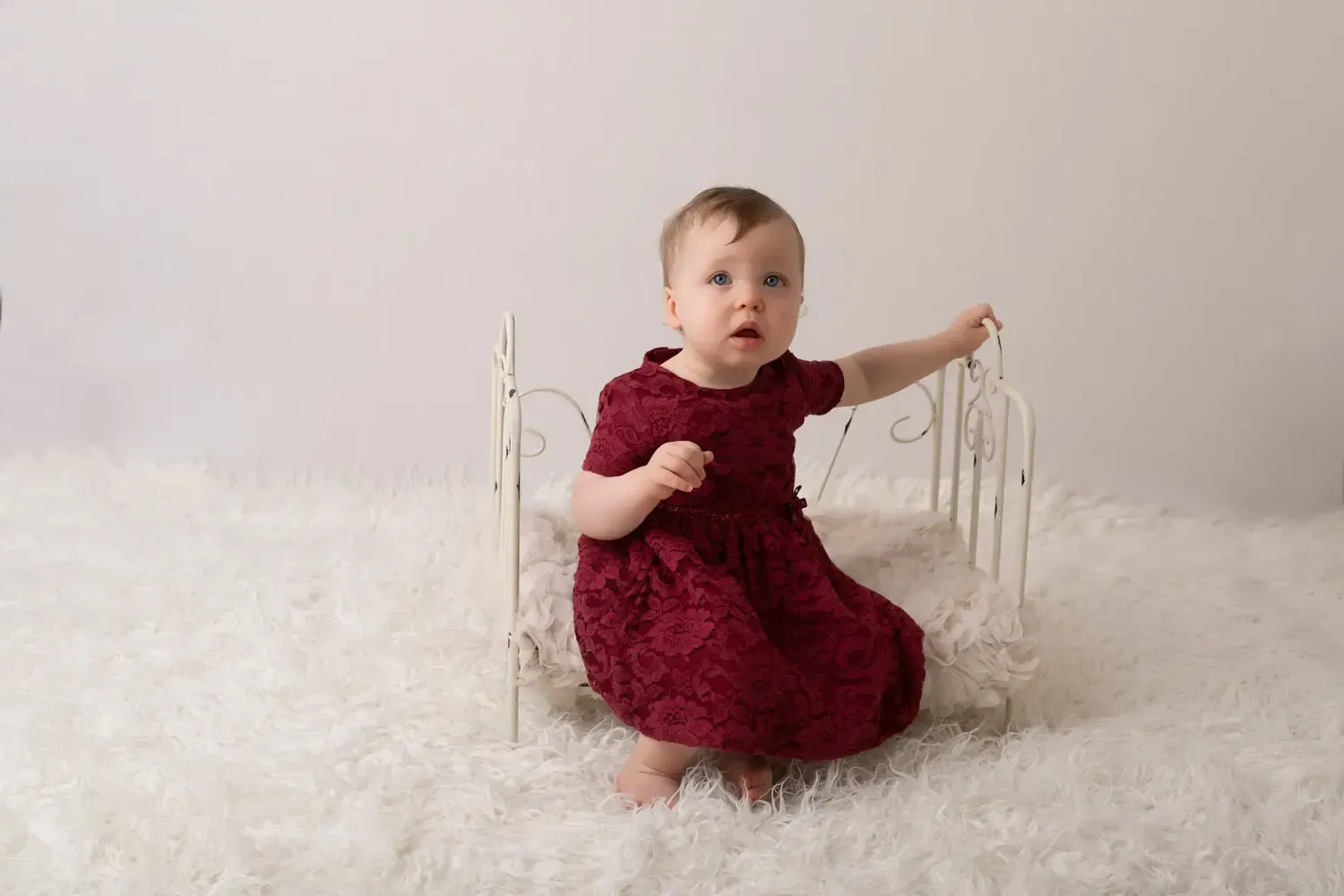 First birthday girl photos in maroon dress sitting on wire bed.