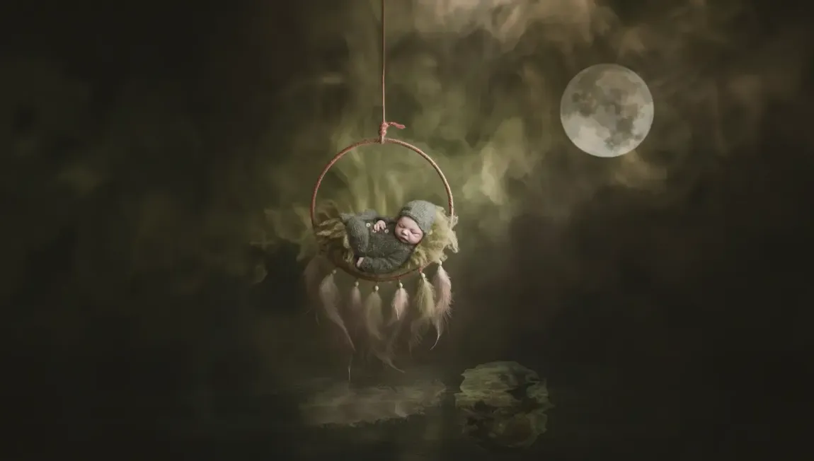 Newborn boy in fuzzy footed romper sleeping under the moon over water.