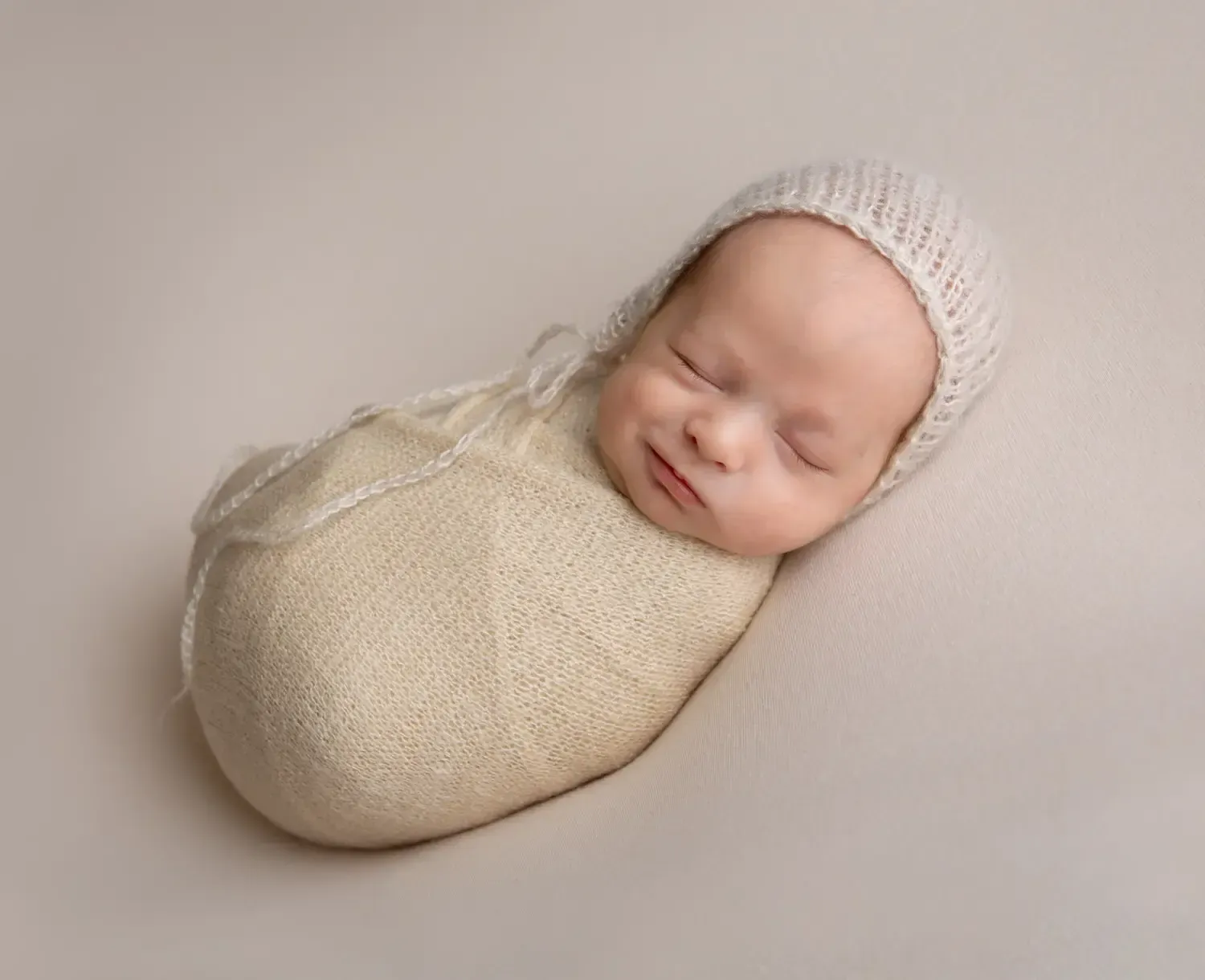 Newborn boy wrapped in taupe with matching bonnet. Smirking.