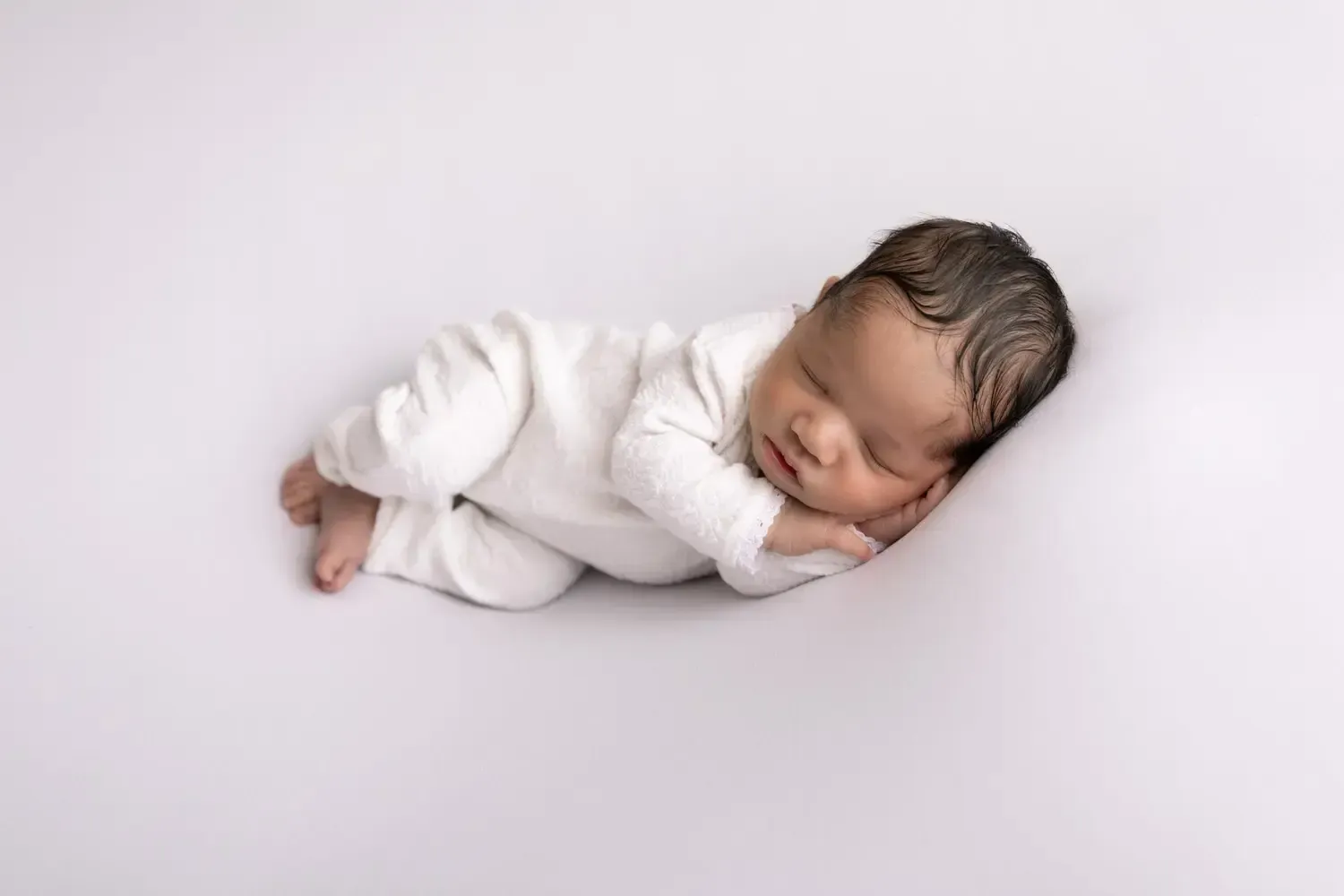 Newborn boy in white romper sleeping on his side.