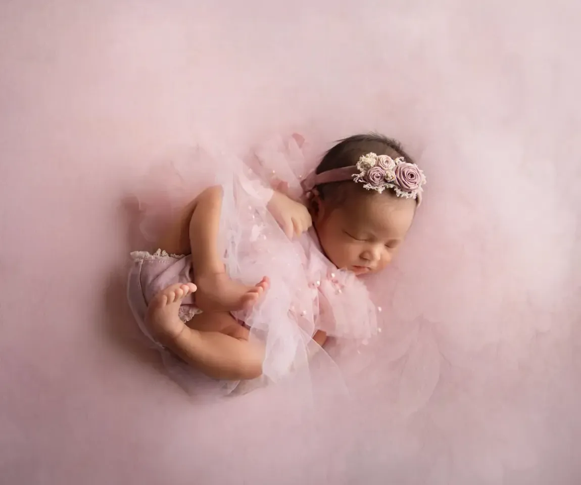 Newborn girl in pink dress. Feet curled up on pink background.