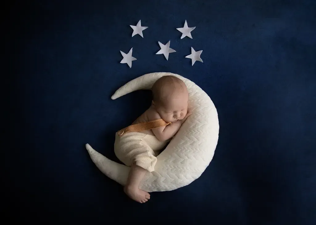 Newborn boy in cream overalls, hugging a moon pillow. stars above. Sleeping.