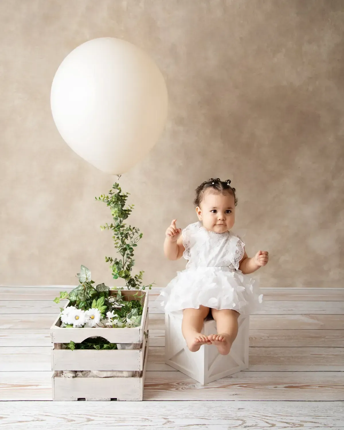 Baby girl siting on white box. One big balloon. White dress.