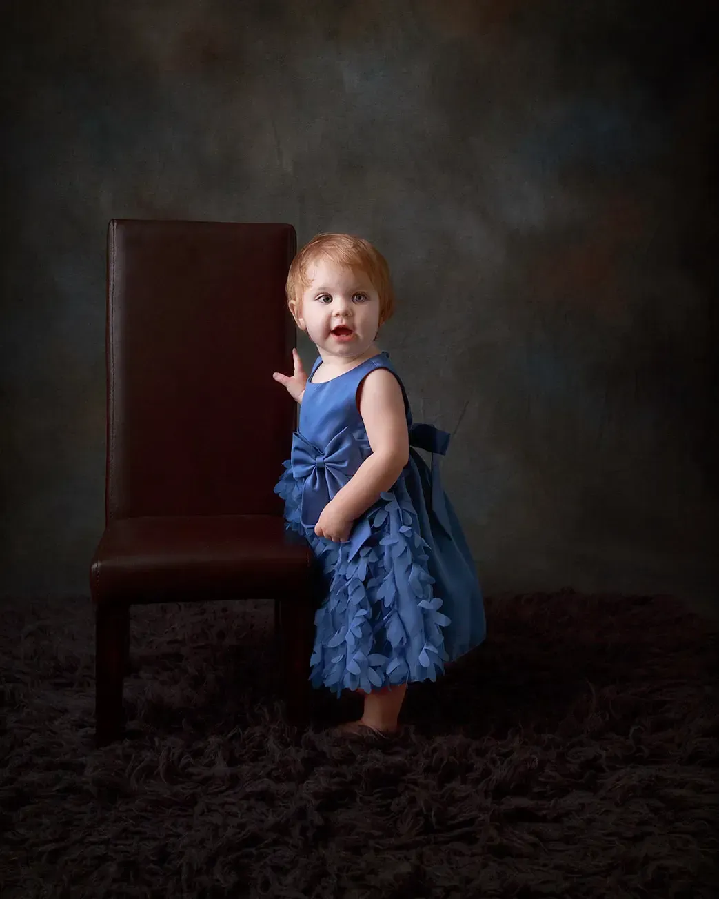 Red headed 8 months 
 girl in blue dress standing near a chair. Dramatic light. 