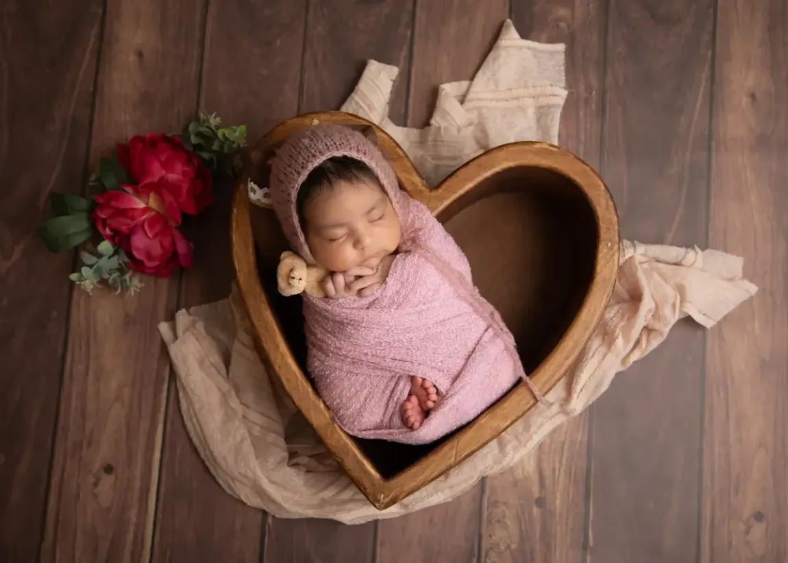 Newborn girl wrapped in pink with pink bonnet. Heart shaped bowl. Holding whitenteddy bear.