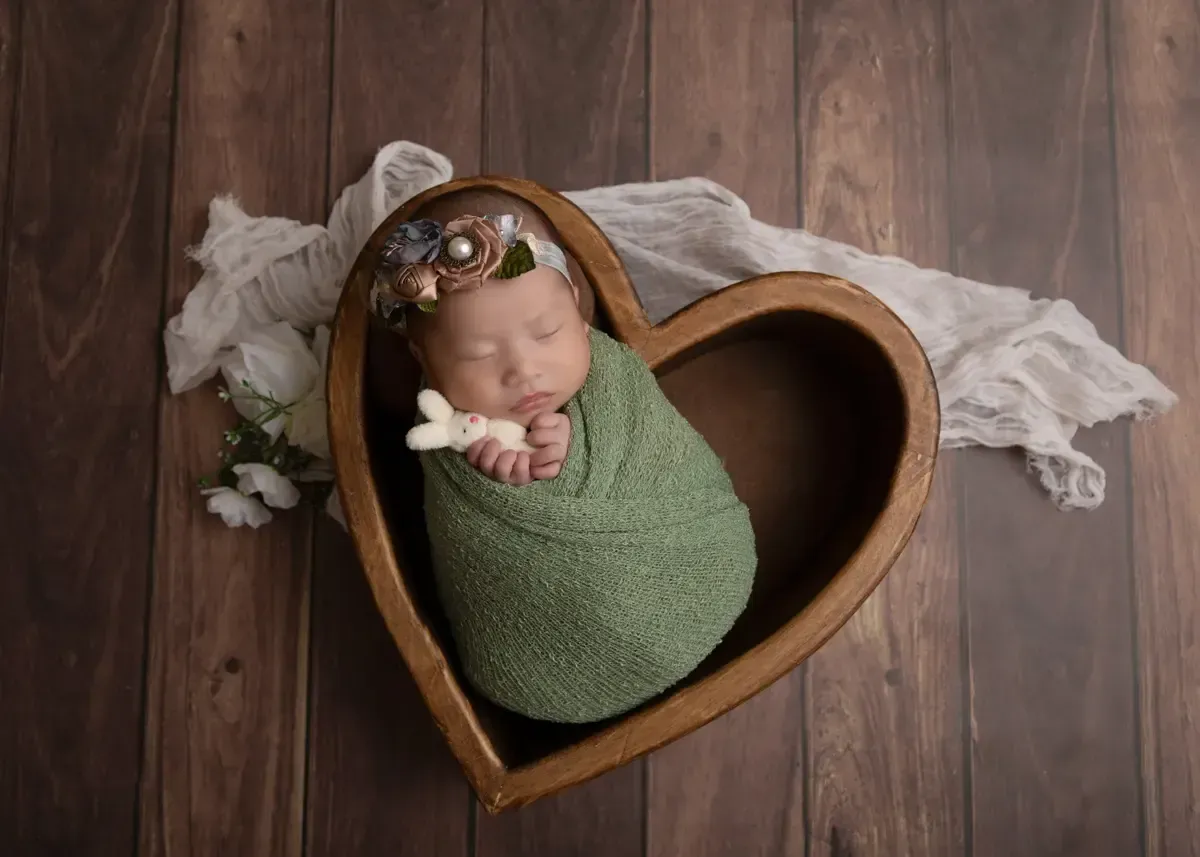 Newborn girl wrapped in green holding stuffie. Placed in wooden heart shaped bowl.
