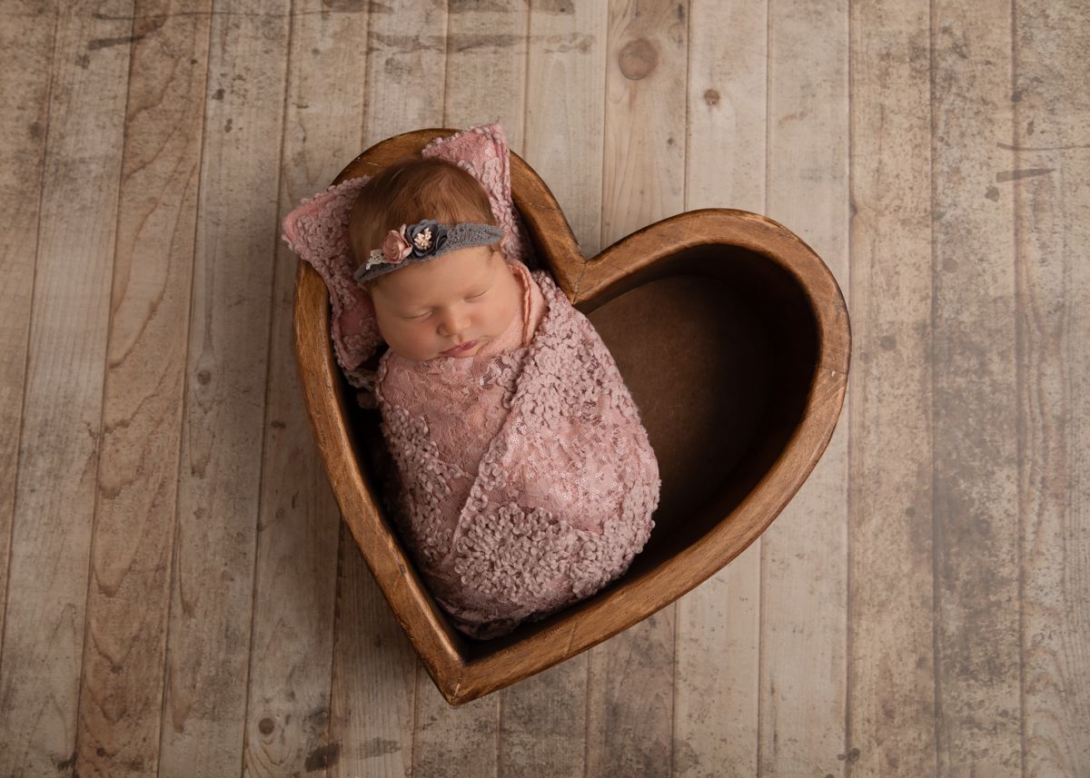 Newborn girl in wooden heart prop wrapped in pink.