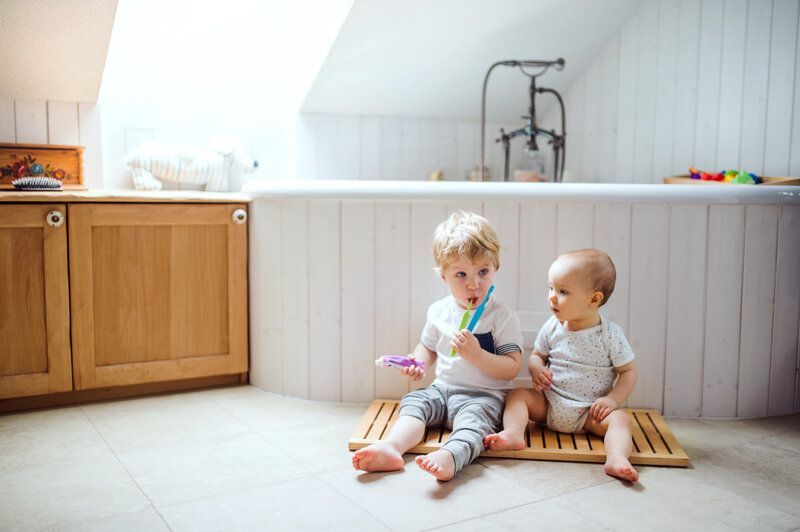 A toddler with his 7 month old sibling brushing his teeth.