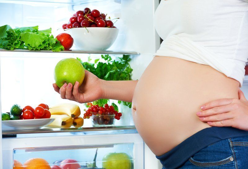 Pregnant woman standing in front of fridge of healty food items.