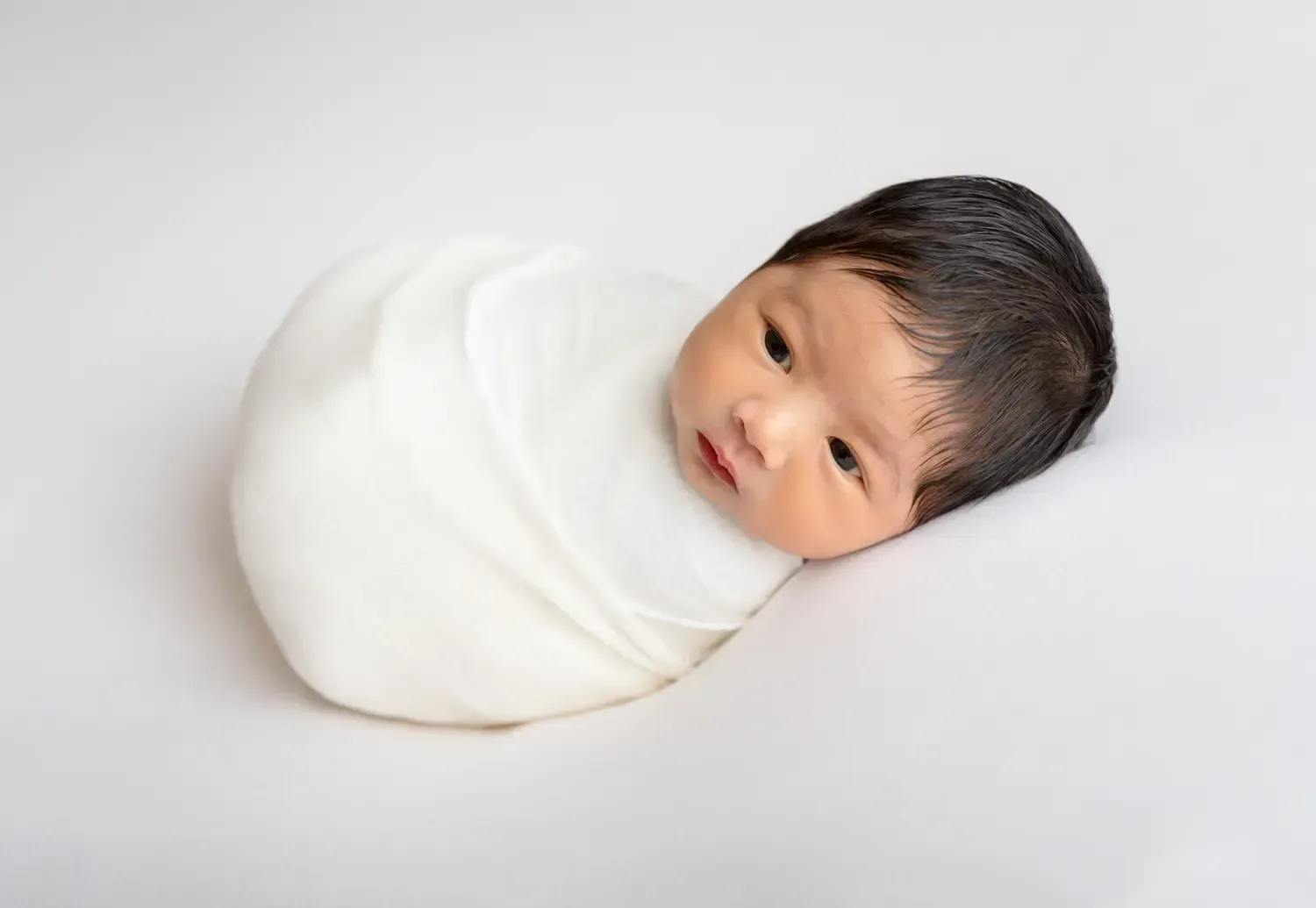 Newborn boy portrait wrapped in white on white backdrop.
