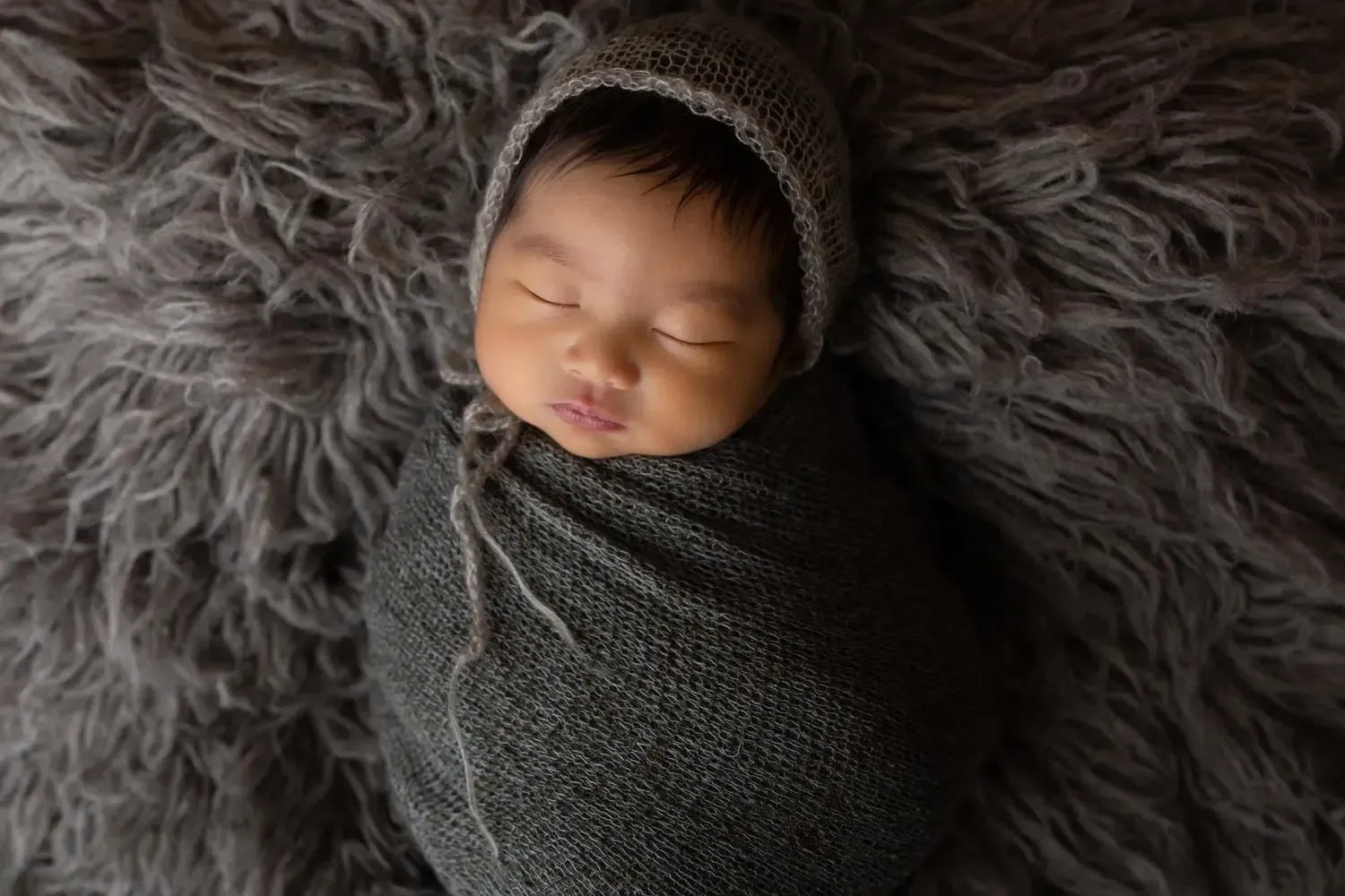 Newborn baby boy in a grey bonnet, wrapped on matching flokati