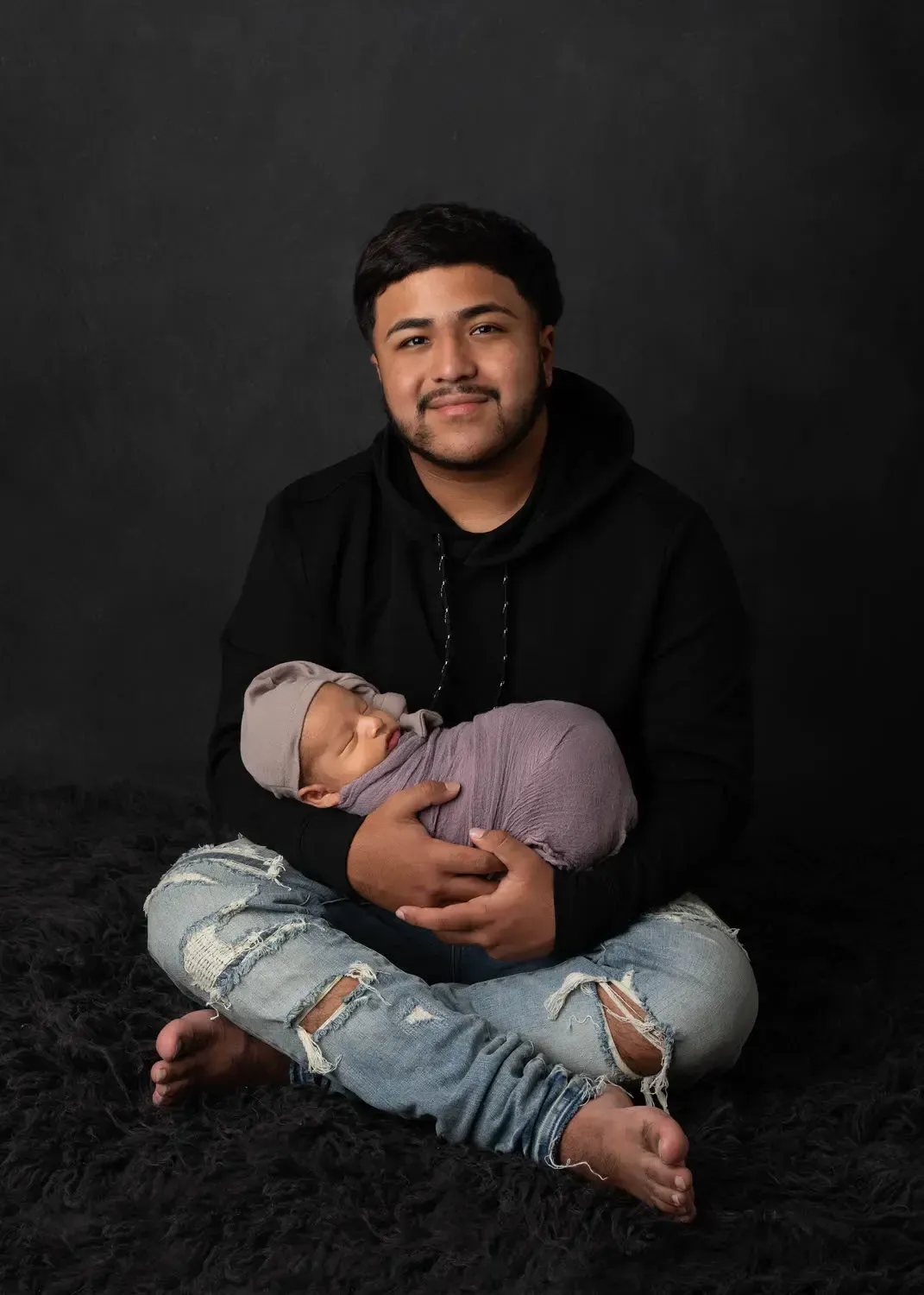 Big Brother with newborn brother. Black shirt, jeans. Sitting crosslegged on floor holding brother in lap.