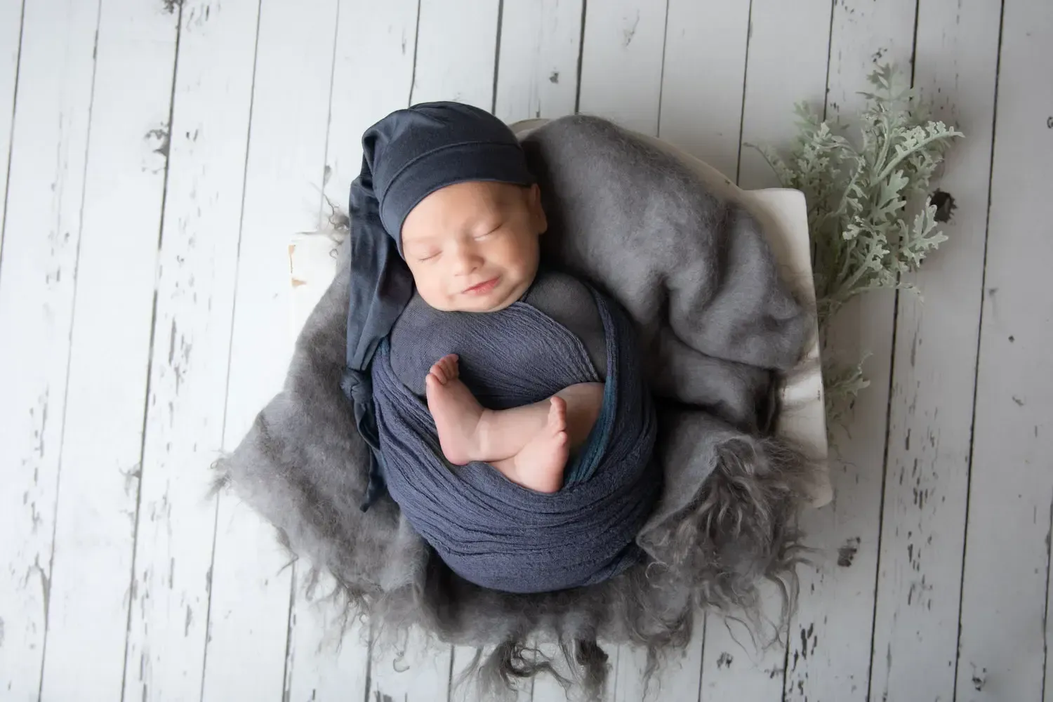 Smilng newborn boy in wooden bowl prop wrapped in blue. Matching sleepy hat.