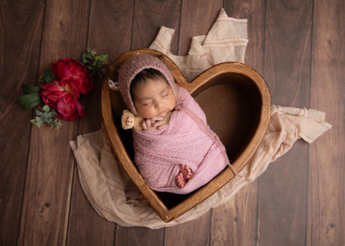 Newborn baby girl wrapped in pink in a hear shaped bowl holding a teddy bear.