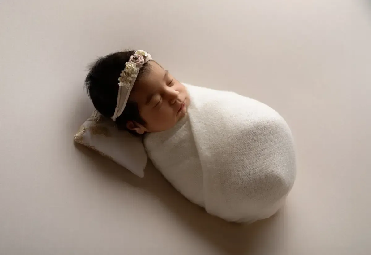 Newborn girl wrapped in white swaddle with floral headband.