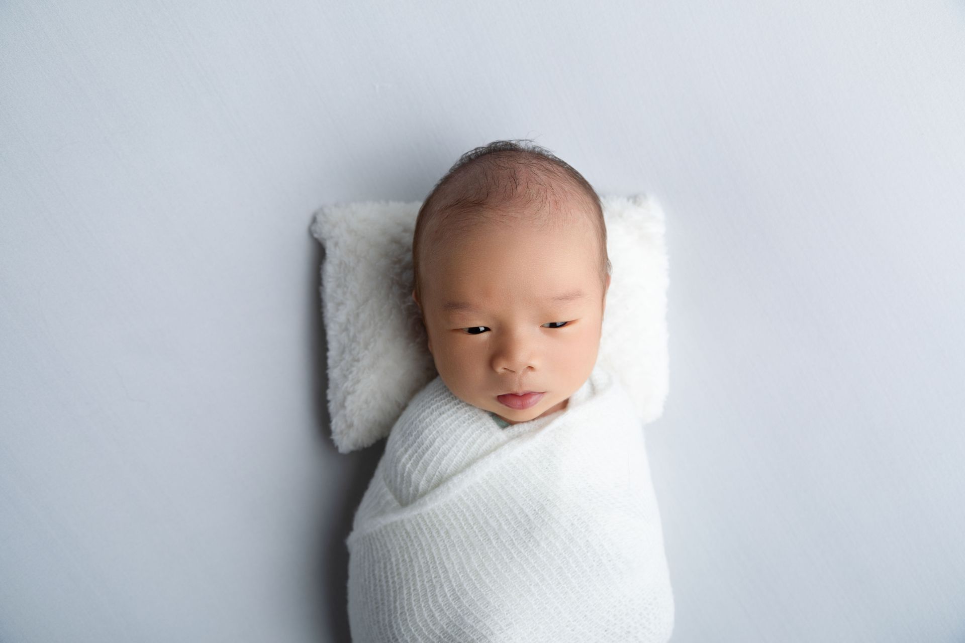 Asian newborn boy in white on white background and pillow. Awake