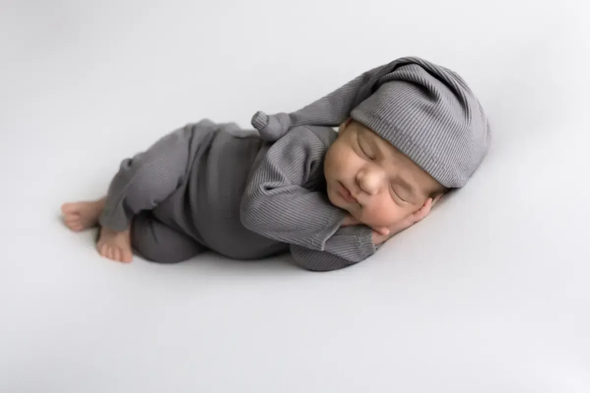 Newborn boy in grey romper with matching sleepy hat sleeping on side.