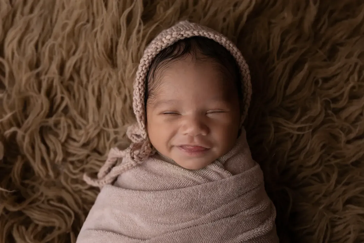 Smiling newborn baby boy wrapped on brown flokati. Matching knitted hat.