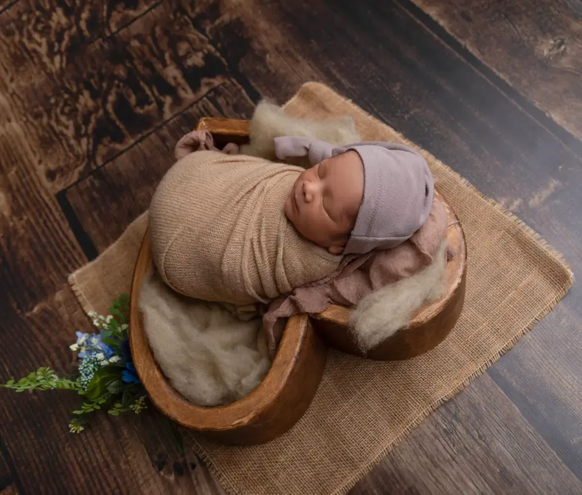 Newborn boy wrapped in tan with matching sleepy hat in heart-shaped wooden bowl.