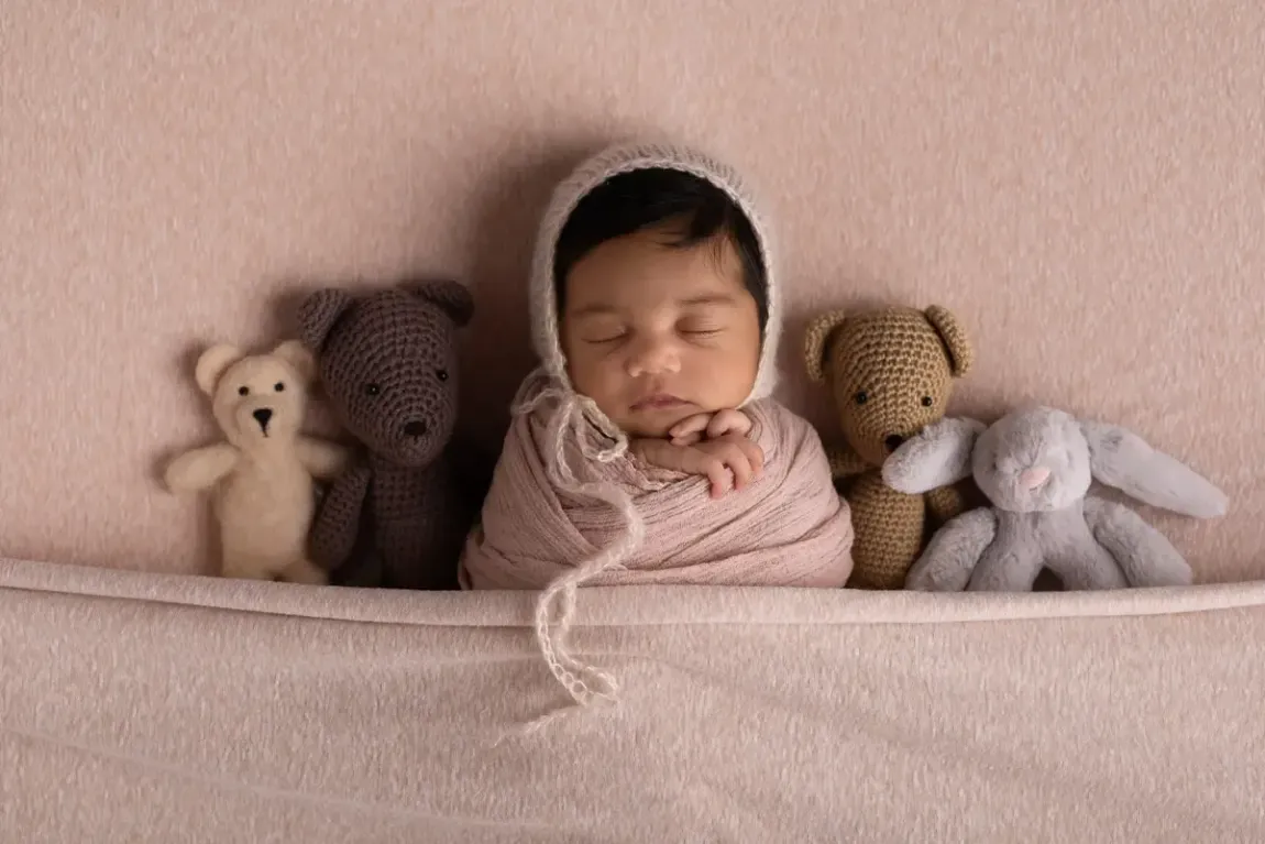 Newborn girl tucked in bed with stuffies. Taupe. Wearing matching bonnet.