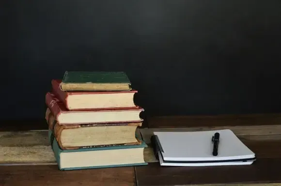 A stack of books on a desk next to a notepad and pen