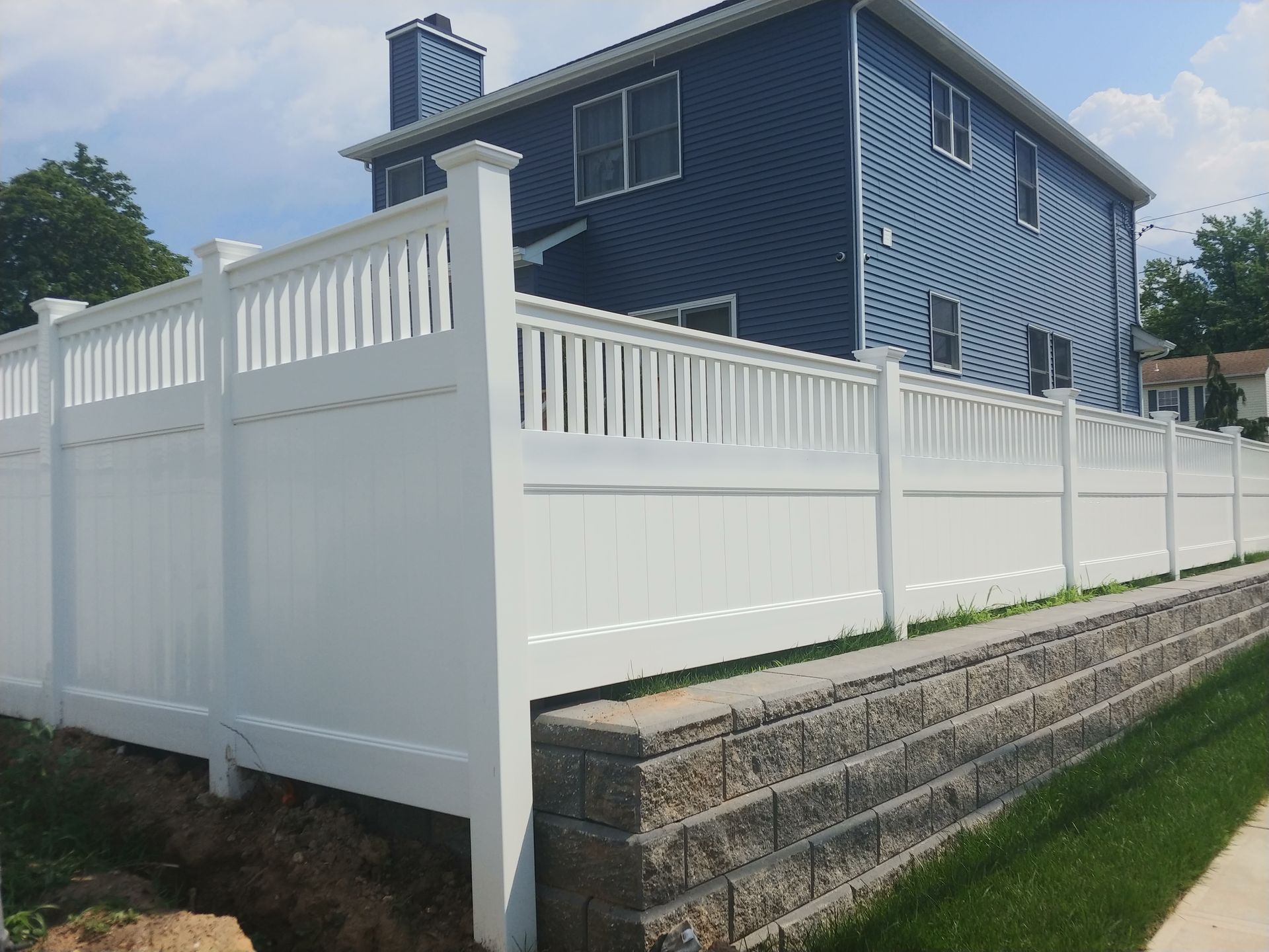 A white fence surrounds a brick wall in front of a blue house.