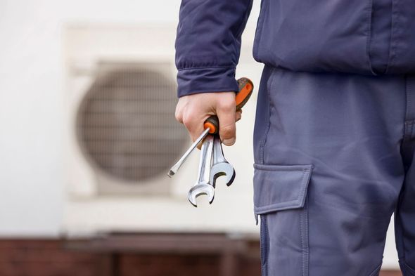 Person in blue workwear holding tools near an air conditioning unit.