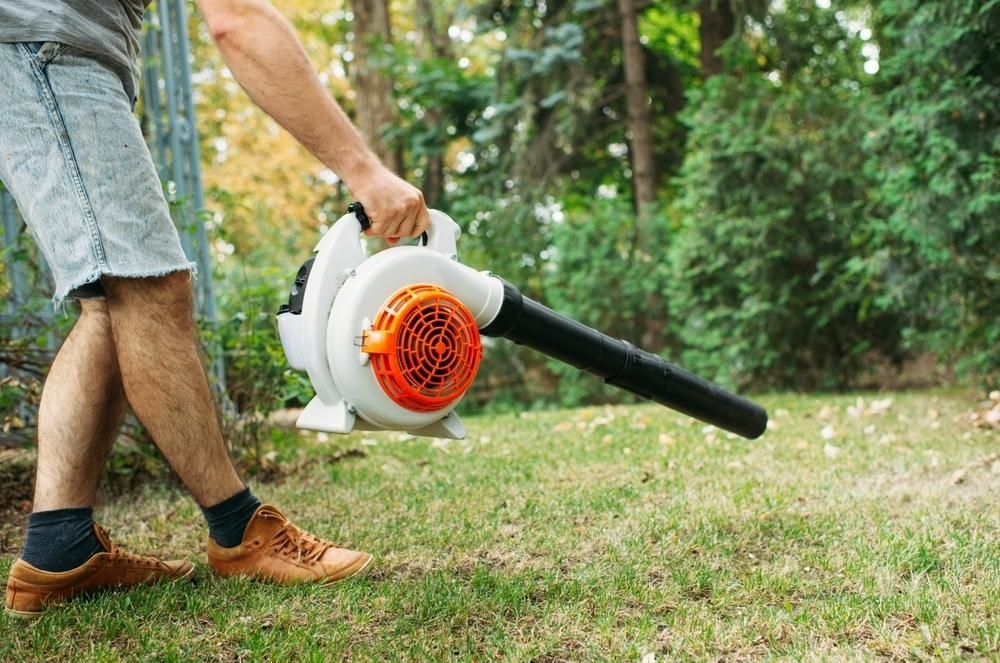 A Man is Using a Leaf Blower to Blow Leaves in a Yard — Urunga Chainsaw and Mower in Urunga, NSW