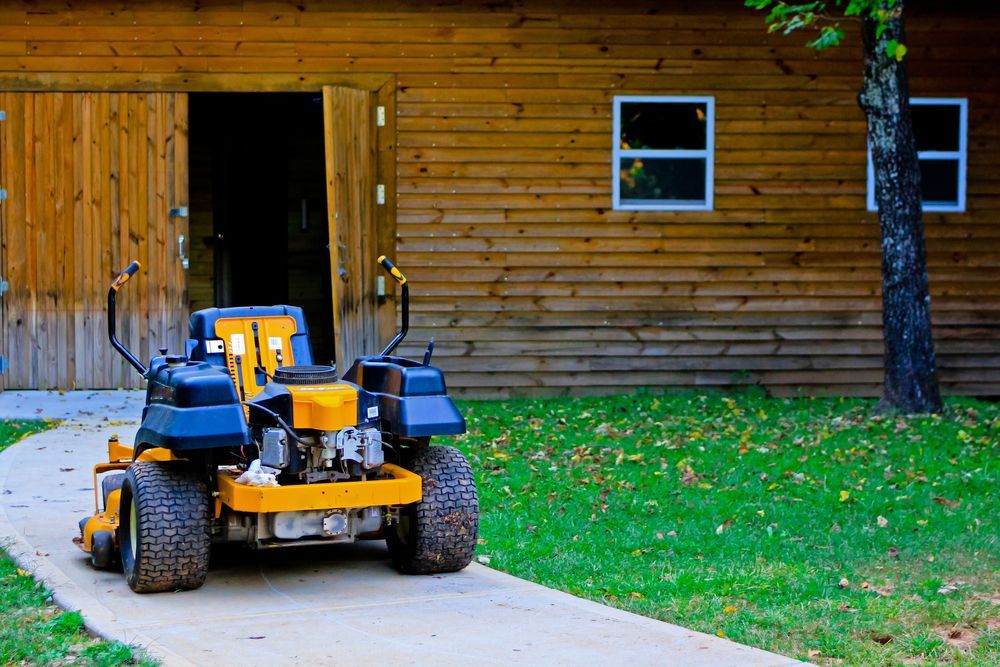 A Yellow and Blue Lawn Mower is Parked in Front of a Wooden House — Urunga Chainsaw and Mower in Mid North Coast, NSW