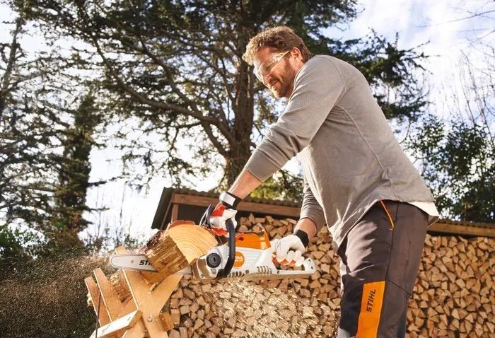 Man using a chainsaw to cut wood outdoors, wearing safety glasses and gloves, sawdust flying.