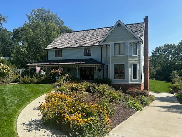 A large white house with a brick chimney is surrounded by flowers and trees.