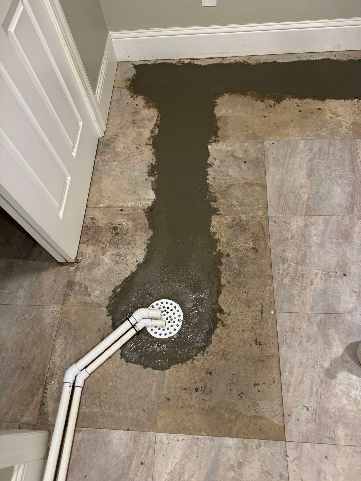 A patch of wet concrete floor repair surrounds a drain, with two white pipes extending from it across the tiled surface.