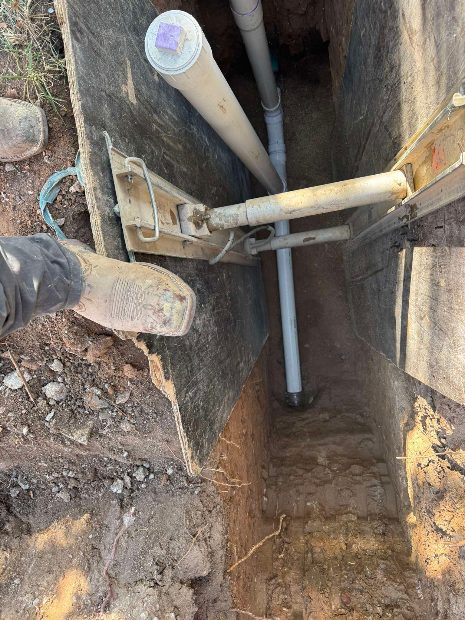 A worker's boot stands at the edge of a deep trench shored up with metal struts, revealing underground PVC pipes.