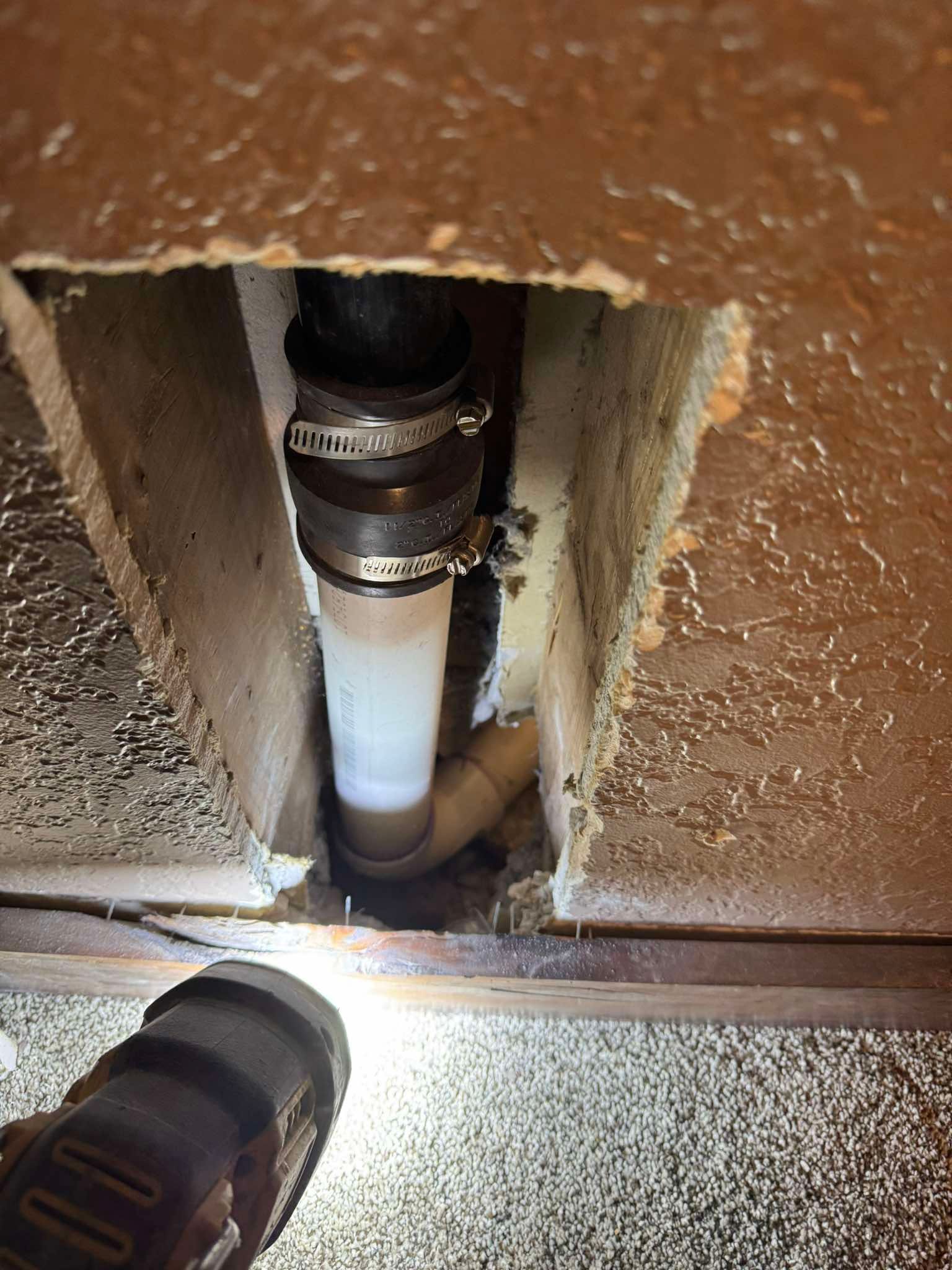 A flashlight illuminates a rubber plumbing coupling connecting a white pipe to a dark pipe inside an open wall section.