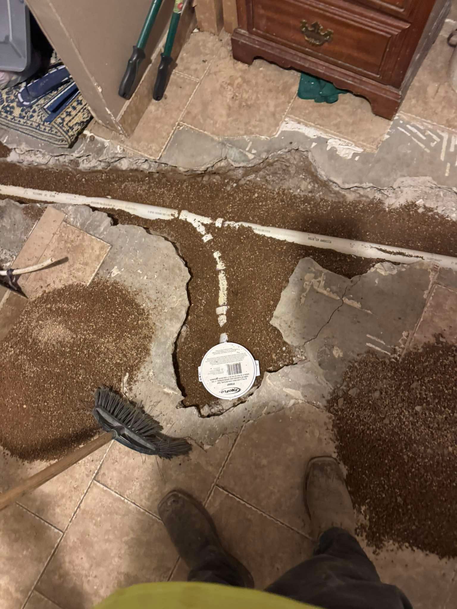A person’s feet stand near a floor trench revealing a white PVC pipe and a cleanout plug amidst loose dirt and debris.