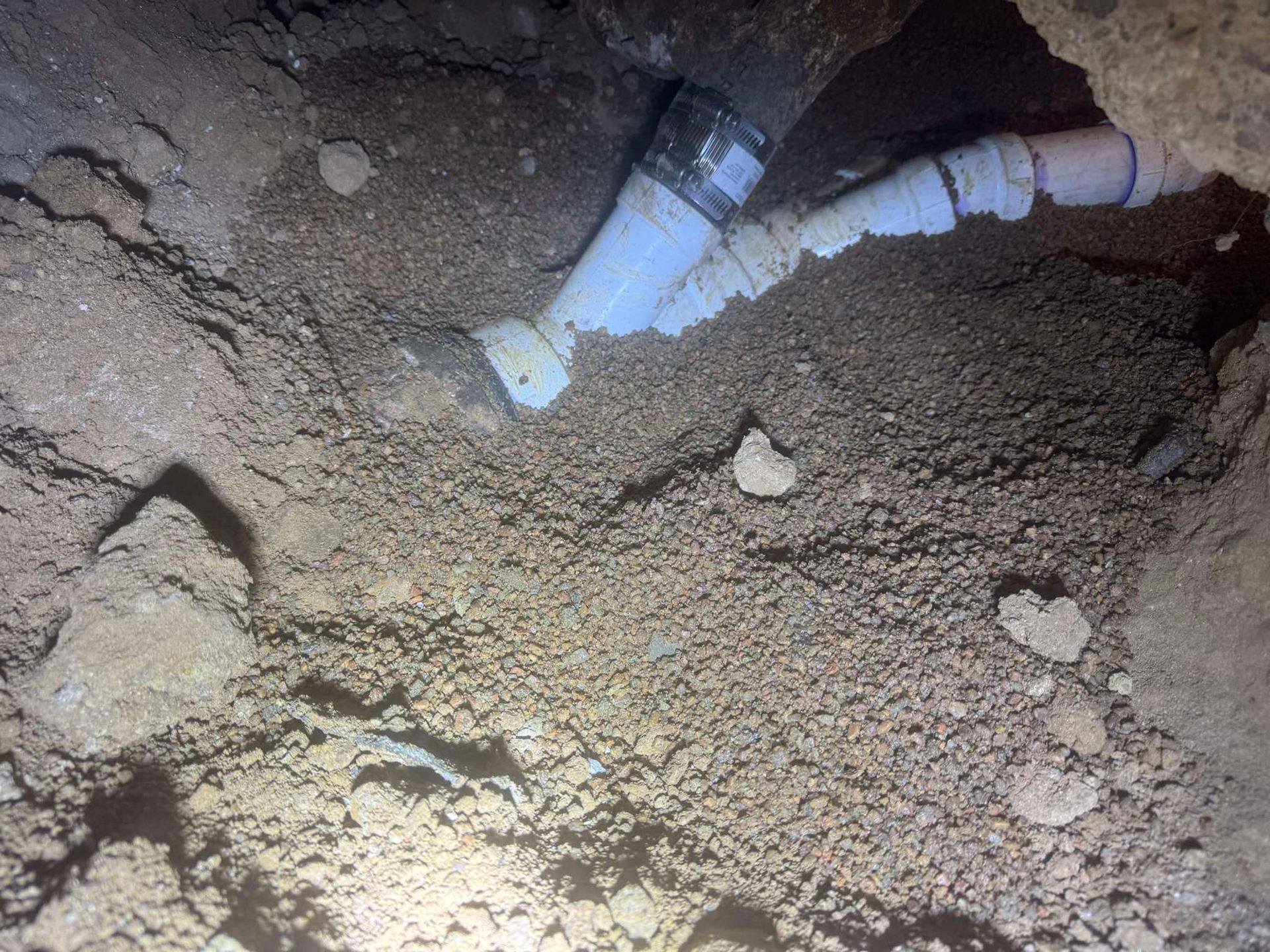 A PVC pipe connection with a wye fitting buried in dirt inside a narrow trench.
