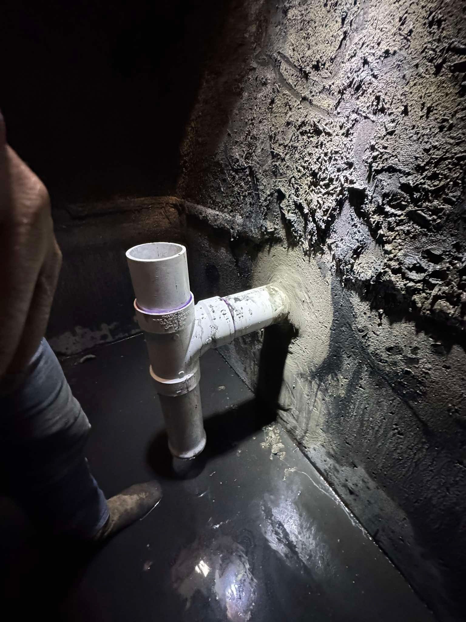 A white PVC pipe assembly with a T-fitting installed against a rough, dark wall in a dimly lit, damp space.