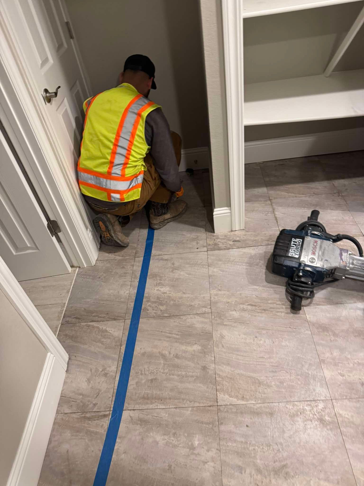 A worker in a high-visibility vest kneels on a tiled floor by a blue tape line, with a demolition hammer nearby.