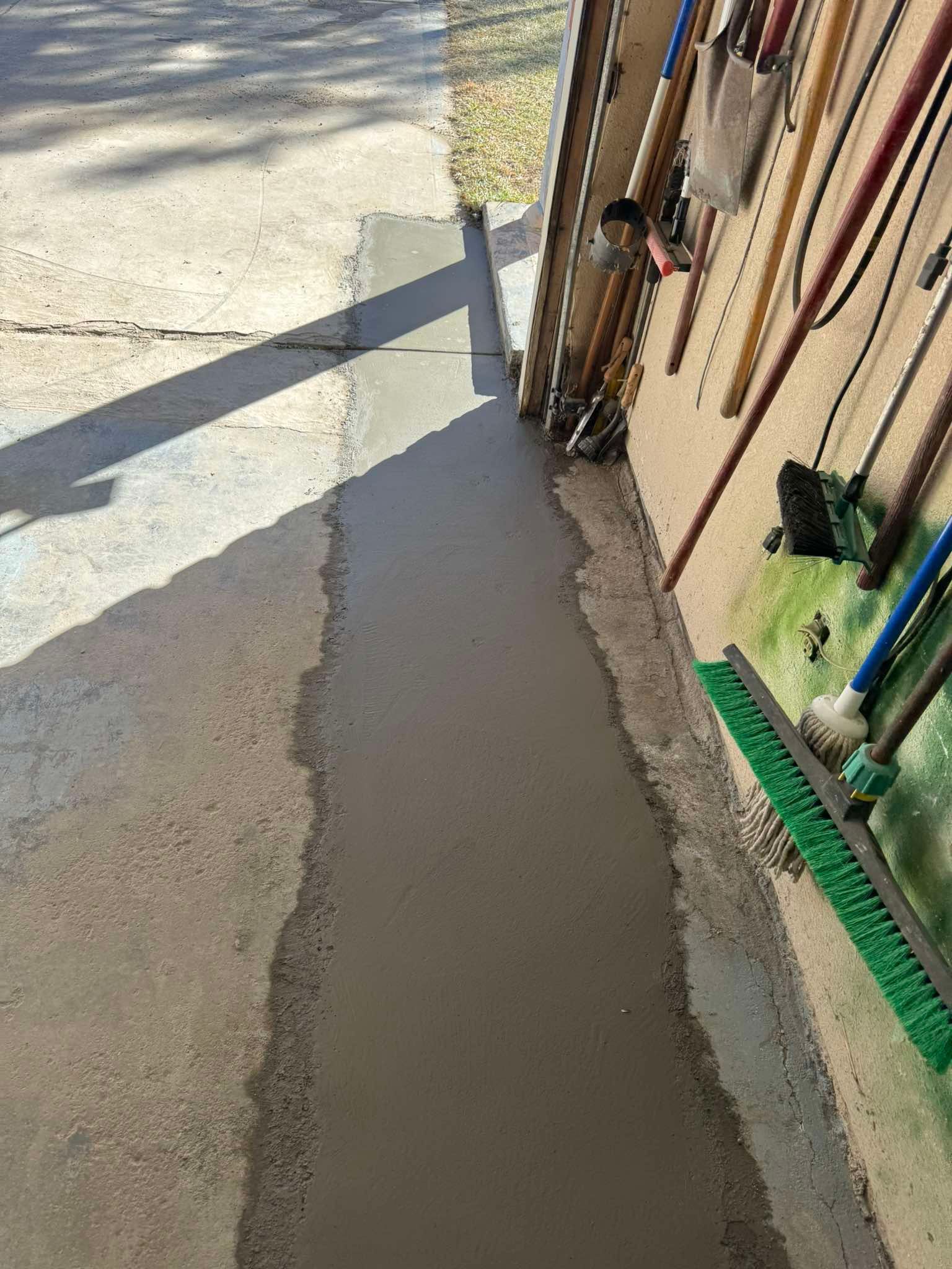 A freshly poured concrete strip along the edge of a garage wall, next to hanging cleaning tools.