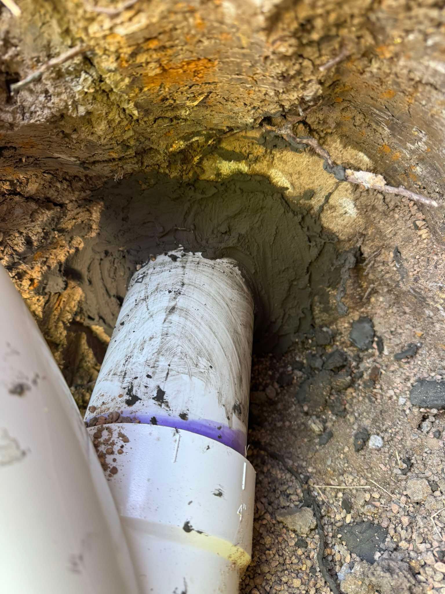 A white PVC pipe enters a hole in the ground filled with dirt and a grey concrete-like sealing material.