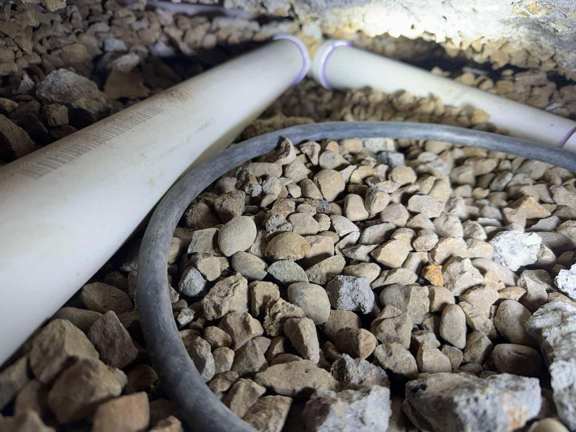 White PVC pipes and a grey circular conduit cable lie amidst a bed of gravel and small rocks.