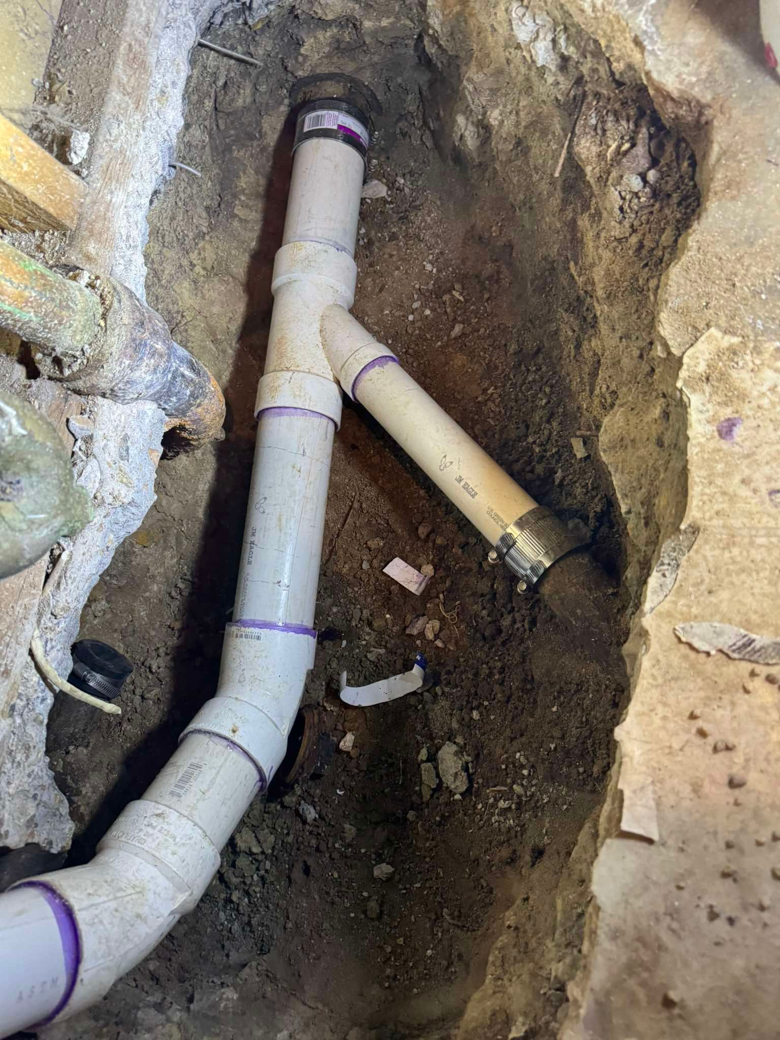 PVC drain pipes connected with a Y-fitting and a metal coupling, installed in a narrow dirt trench beneath a floor.