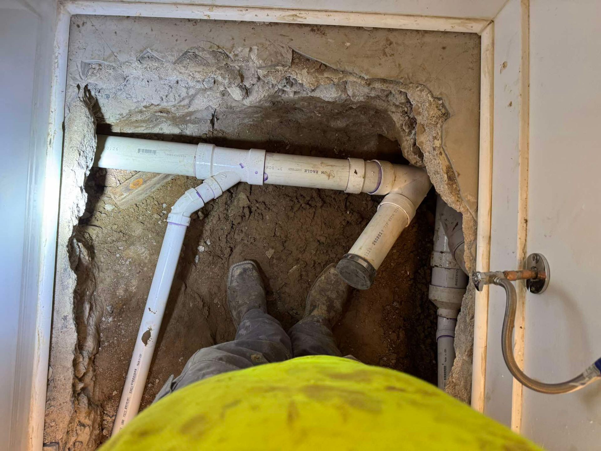 A person in a yellow safety vest stands over an excavated trench in a concrete floor containing exposed PVC plumbing pipes.