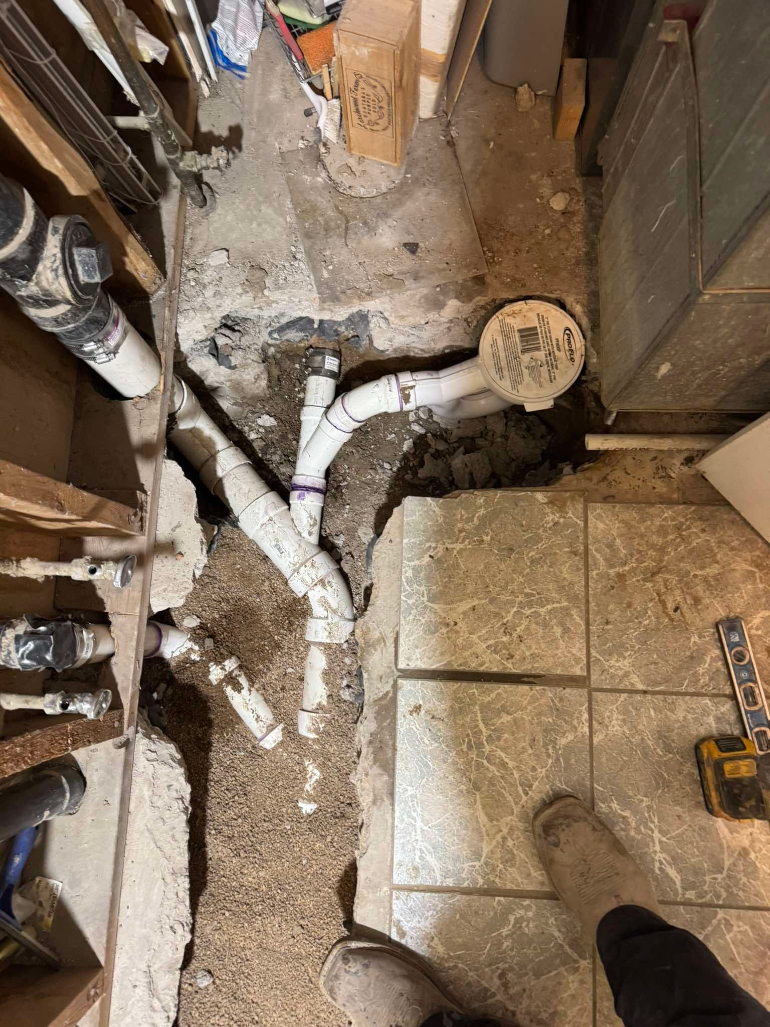Construction site showing PVC pipes connecting to a white circular drain cover in a dirt-filled trench near tile flooring.