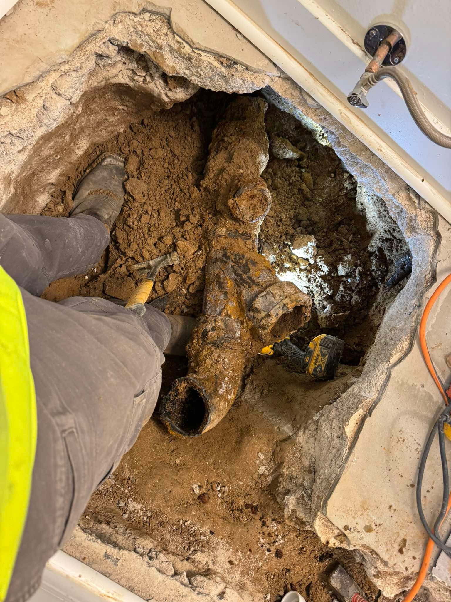 A construction worker stands in a floor cutout, exposing old, corroded plumbing pipes buried in the dirt.